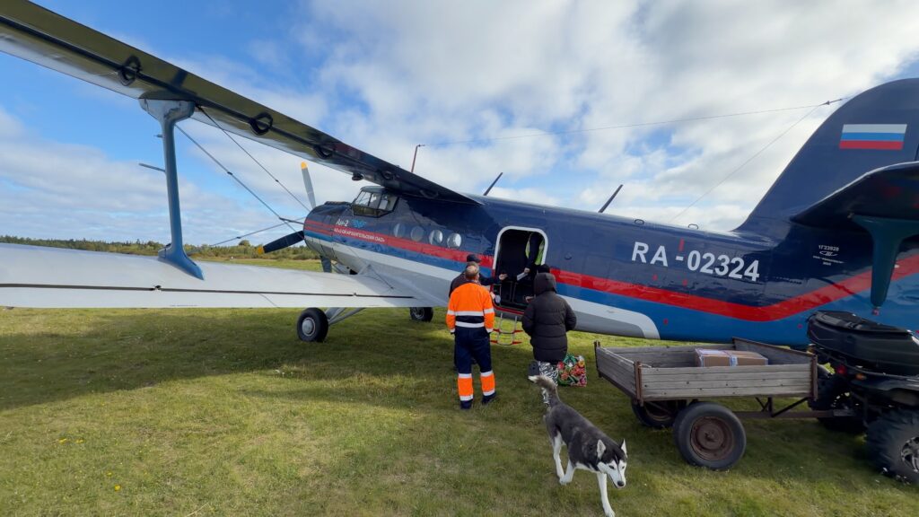 Ан-2ТП RA-02324 в сине-красной ливрее на площадке Пертоминска, багаж из открытой двери грузят в прицеп квадроцикла, рядом хаски