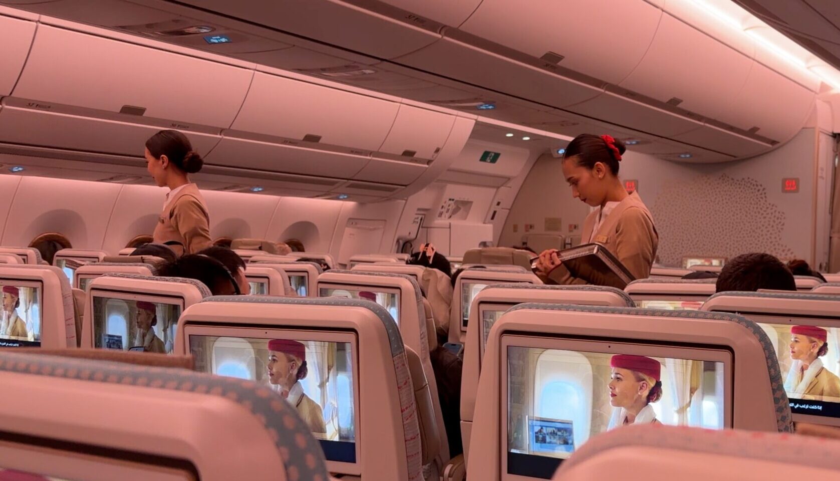 Бортпроводницы Emirates в салоне эконом-класса Airbus A350 с розовой подсветкой, одна из них с меню в руках обходит пассажиров, на экранах в спинках кресел идёт видео авиакомпании